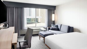 Habitación de hotel con cama, escritorio, sillón, sofá y un amplio ventanal con cortinas.