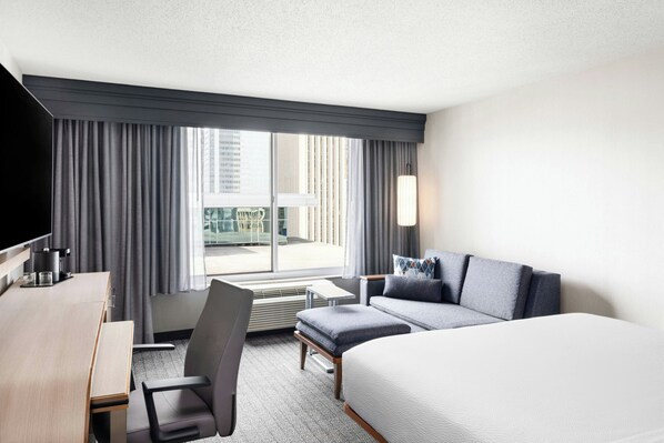 Une chambre d’hôtel avec un lit, un bureau, une chaise, un canapé et une grande fenêtre avec des rideaux.