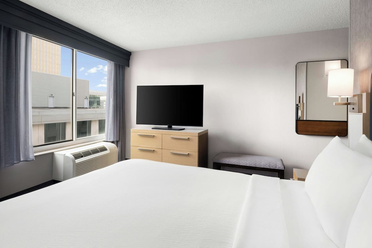 Une chambre d’hôtel avec un grand lit, un téléviseur à écran plat posé sur une commode en bois, une petite table et une fenêtre avec des rideaux.