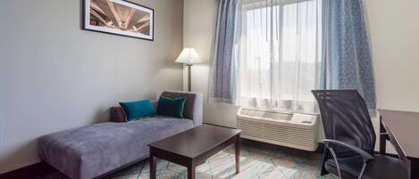 A hotel room with a sofa, a small table, a desk, a chair, a lamp, and a window with curtains.