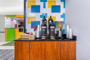 Une petite cuisine équipée d’une machine à café, d’un mixeur et d’un distributeur de café. On y trouve un meuble en bois à deux portes et un plan de travail sur lequel sont disposés divers objets.