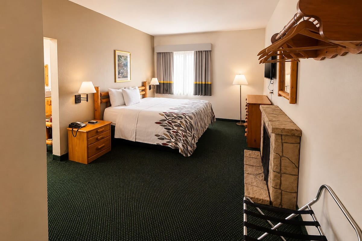 A hotel room with a bed, bedside lamps, a telephone, a small wooden nightstand, a framed picture on the wall, and a stone fireplace.
