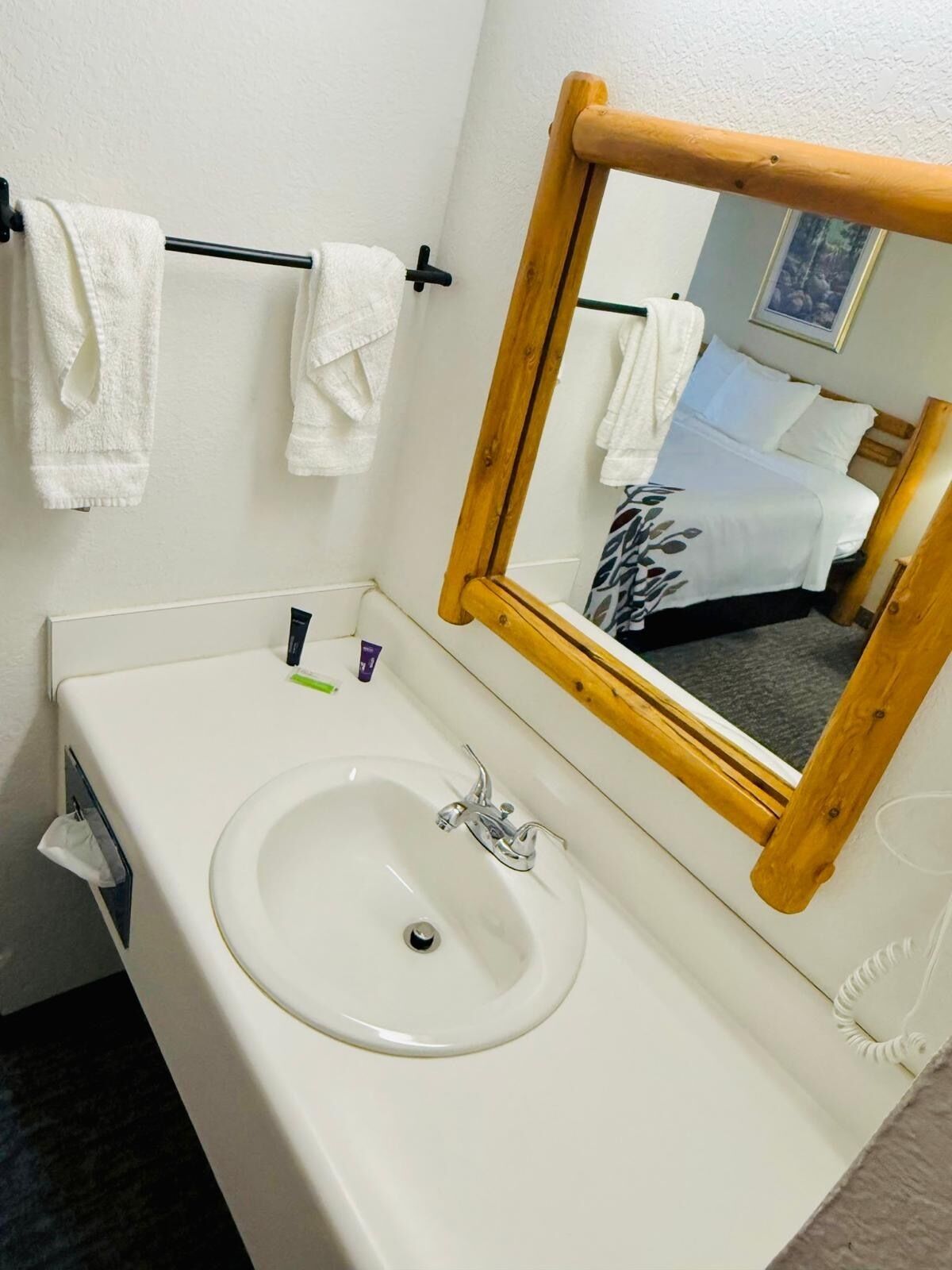 Une salle de bain avec un lavabo blanc, un miroir avec un cadre en bois et deux serviettes suspendues à un porte-serviettes noir.
