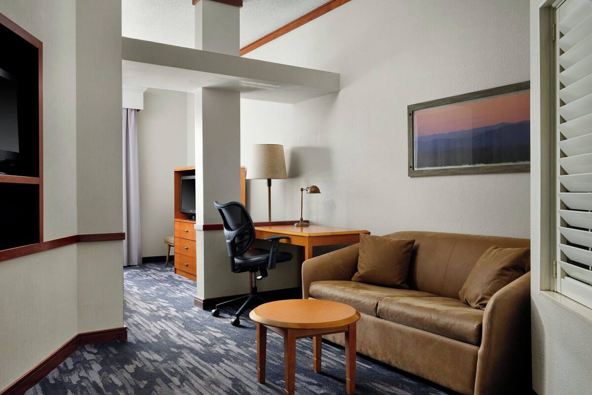 Une chambre d’hôtel avec un canapé en cuir marron, une table d’appoint en bois, un bureau avec une lampe, un fauteuil de bureau noir et un tableau encadré accroché au mur.