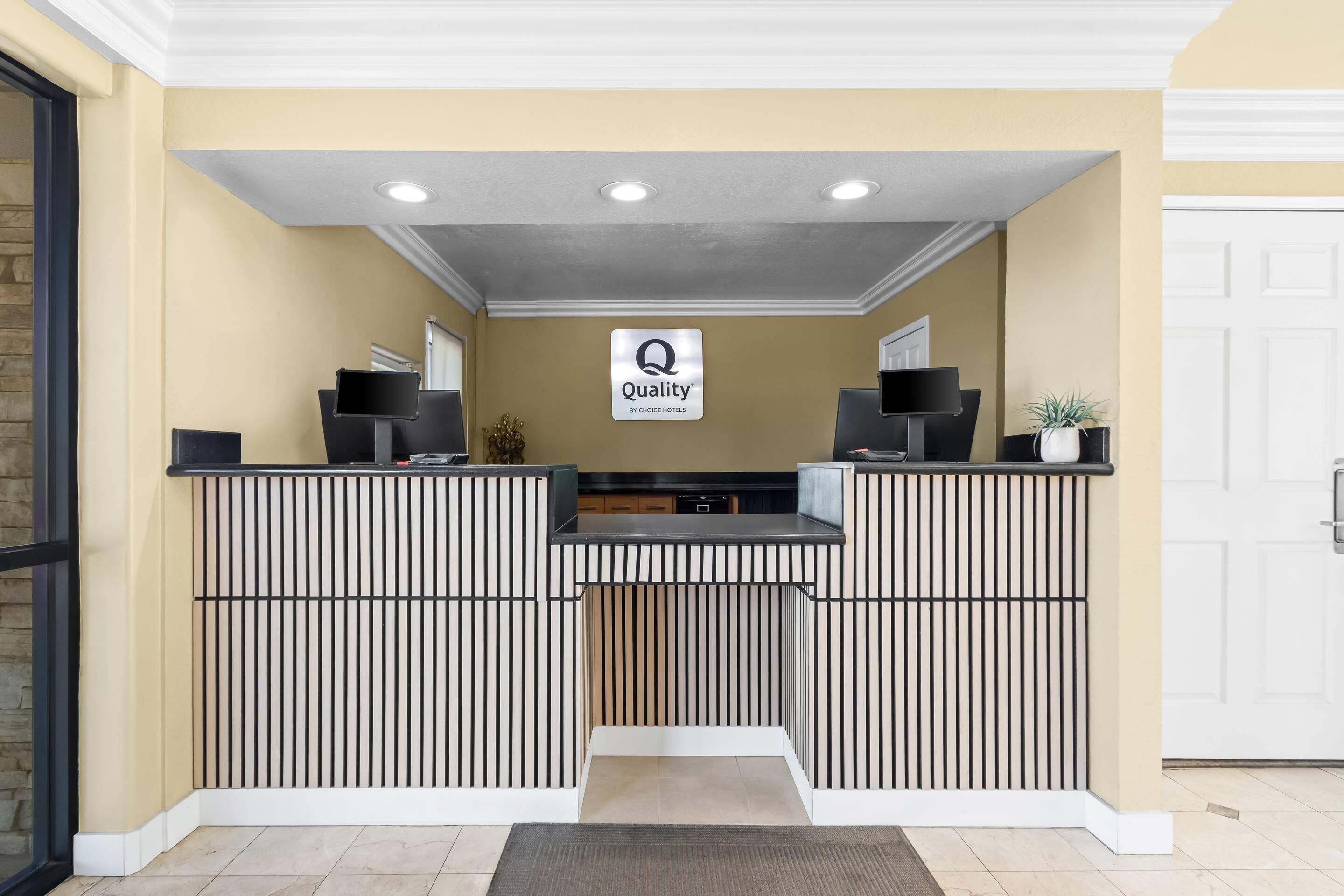 Un mostrador de recepción con un letrero que dice 'Quality by Choice Hotels', un televisor con marco negro y una planta en maceta.