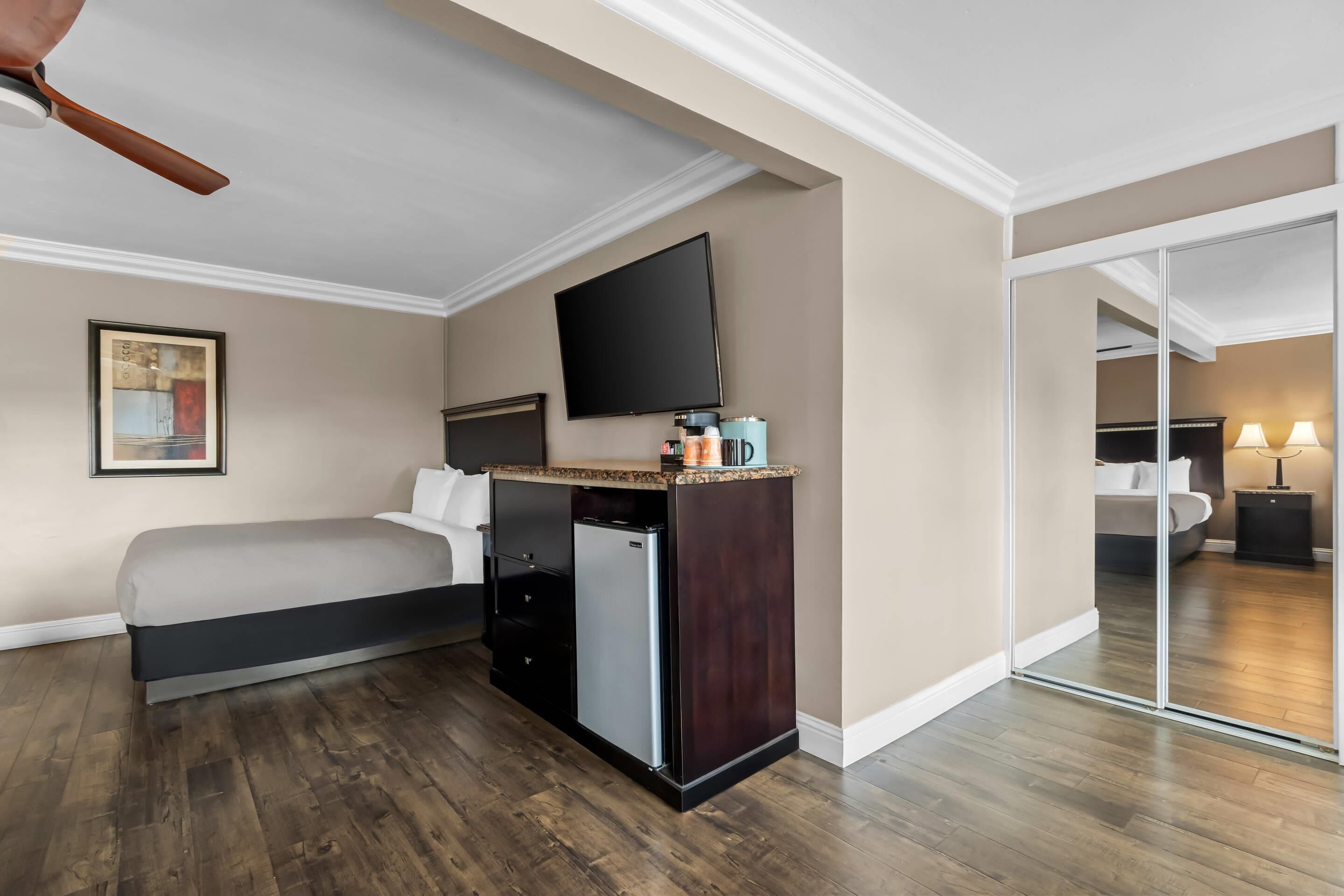 A hotel room with a bed, a flat-screen TV, a small kitchenette, and a sliding door to a bathroom.