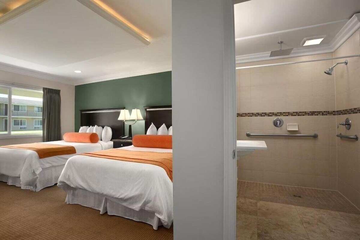 Une chambre d’hôtel avec deux lits, une table de chevet, une lampe, une salle de bain avec douche et vue sur un bâtiment à l’extérieur.