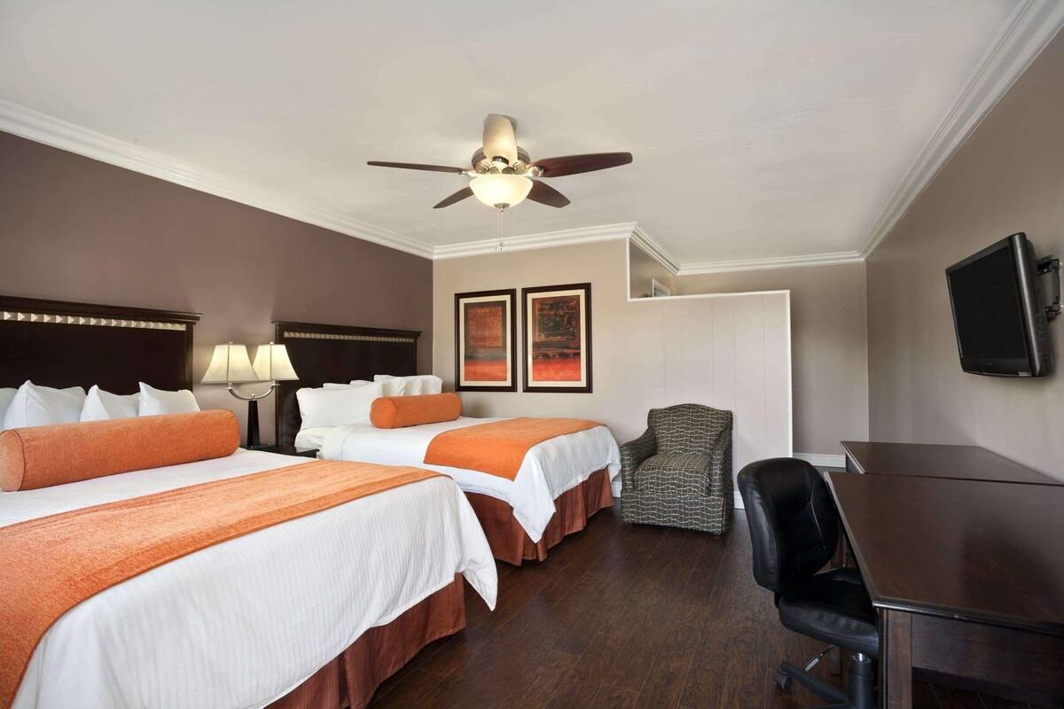 A hotel room with two beds, a desk, a chair, a ceiling fan, and two framed pictures on the wall.