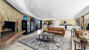 Een hotel lobby met een bruine leren bank, een bijzettafeltje, een bloemenkleed, een koelkast voor Monster energy drink en een keuken in de achtergrond.
