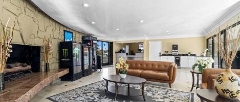 A hotel lobby with a brown leather sofa, a coffee table, a floral rug, a Monster energy drink fridge, and a kitchen area in the background.