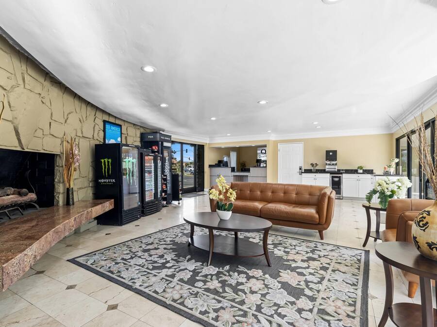A hotel lobby with a brown leather sofa, a coffee table, a floral rug, a Monster energy drink fridge, and a kitchen area in the background.