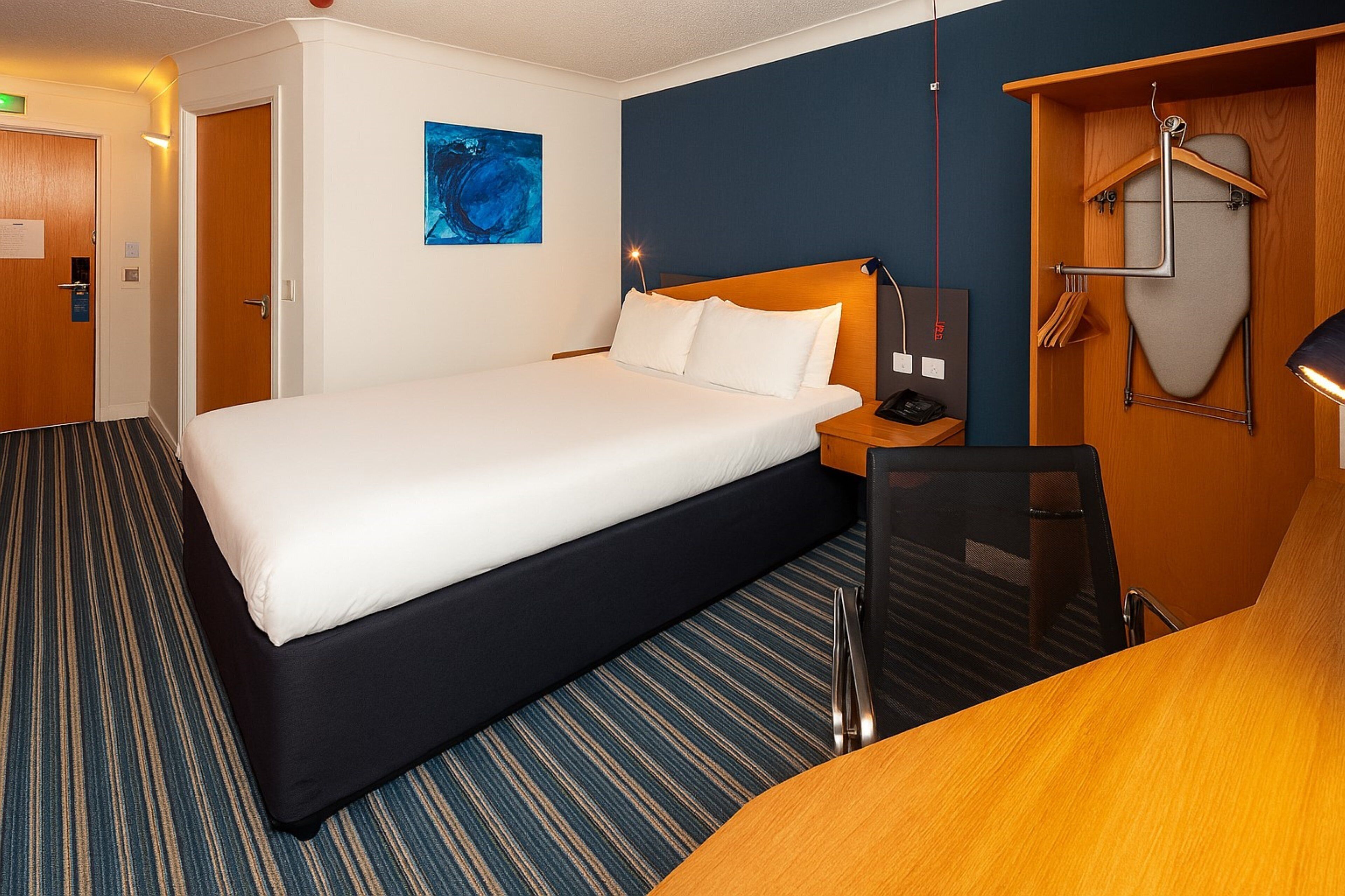 Une chambre d’hôtel avec un lit, un bureau, une chaise, un porte-manteau et un tableau au mur.