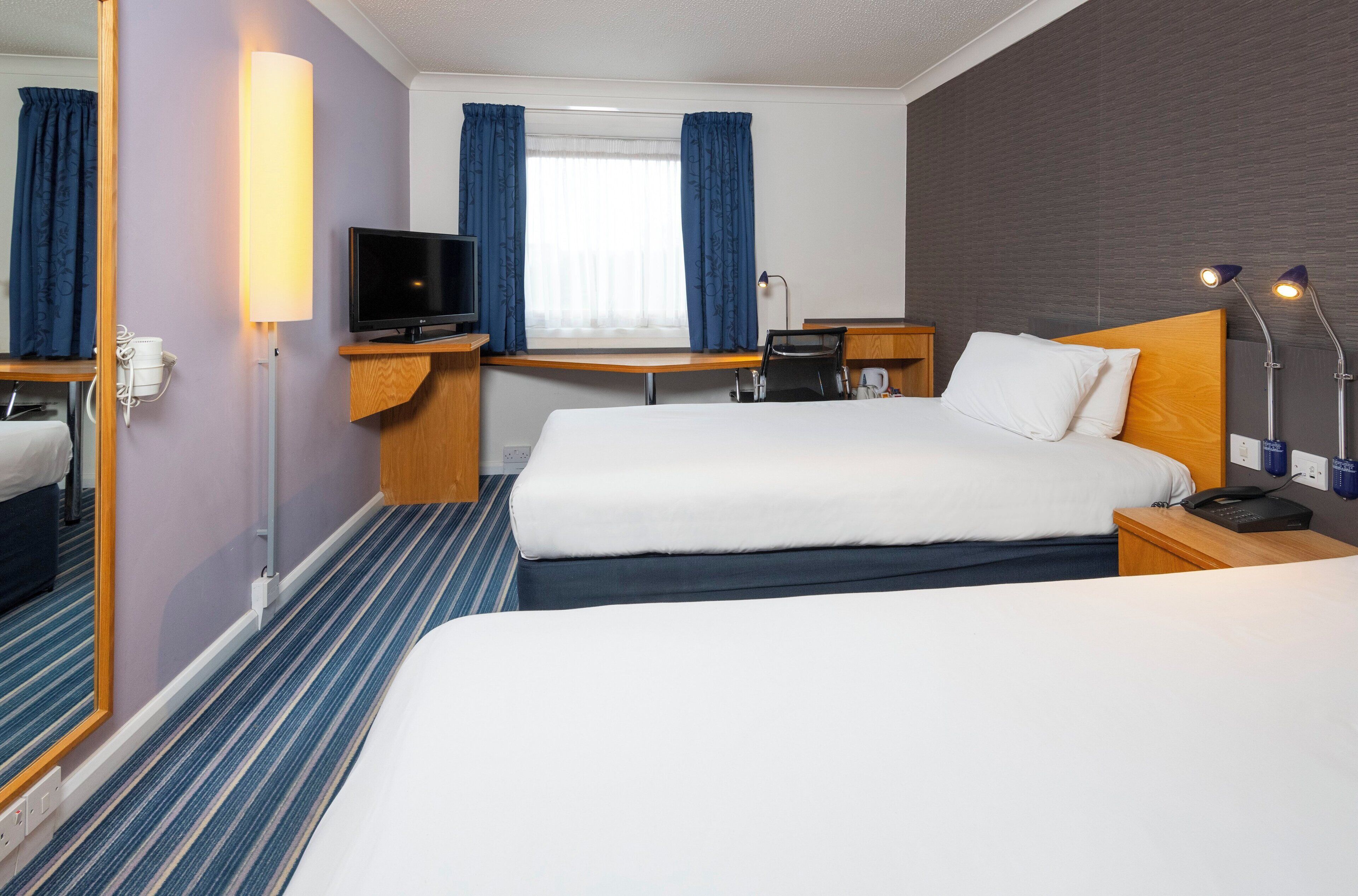 A hotel room with two beds, a desk, a television, and a window with curtains.
