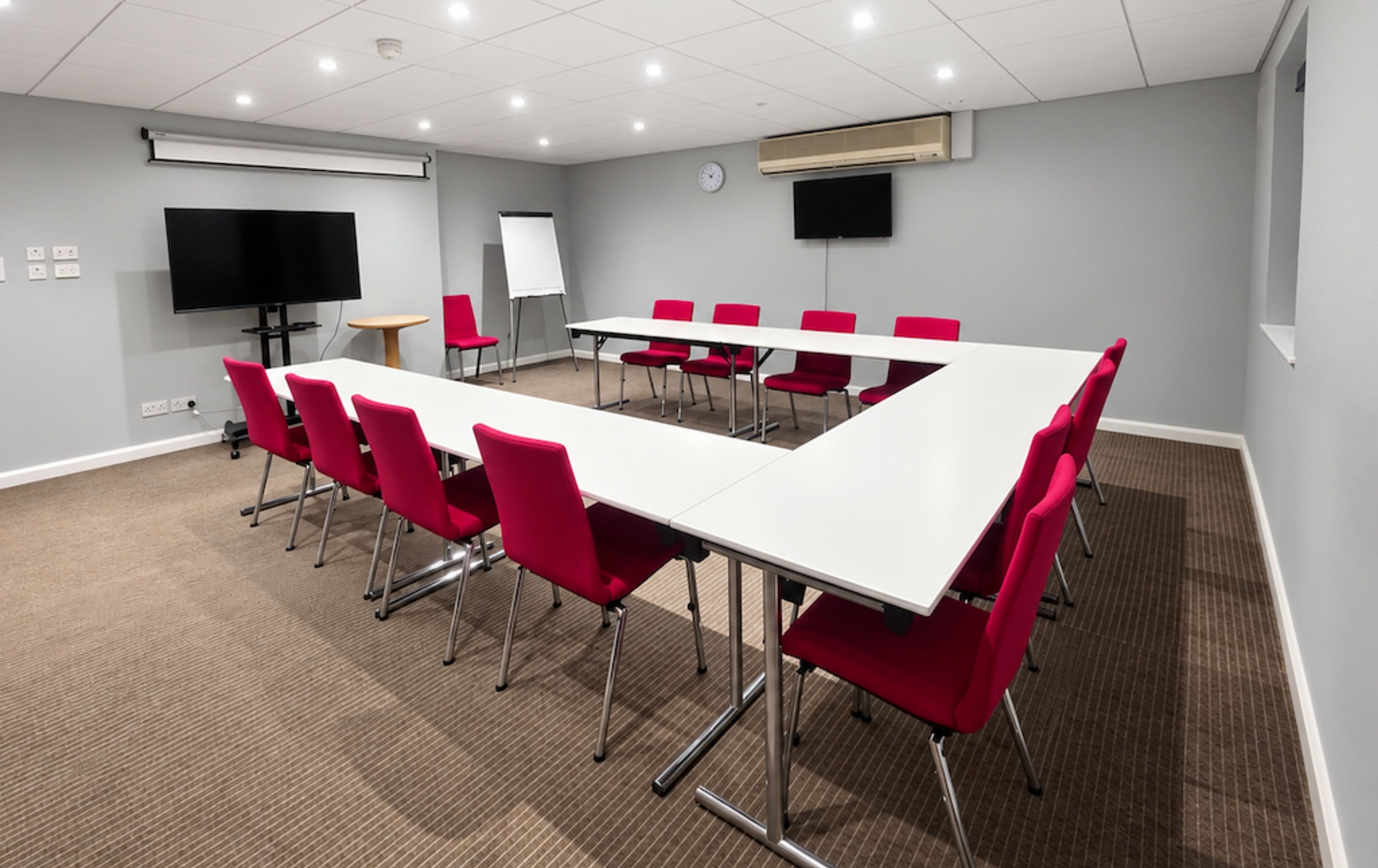 A conference room with a long table, red chairs, a whiteboard, a clock, and a flat-screen TV.
