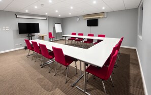 Une salle de conférence avec une longue table, des chaises rouges, un tableau blanc, une horloge et un téléviseur à écran plat.