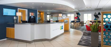 A hotel reception area with a counter, a clock, and a seating area with tables and chairs.