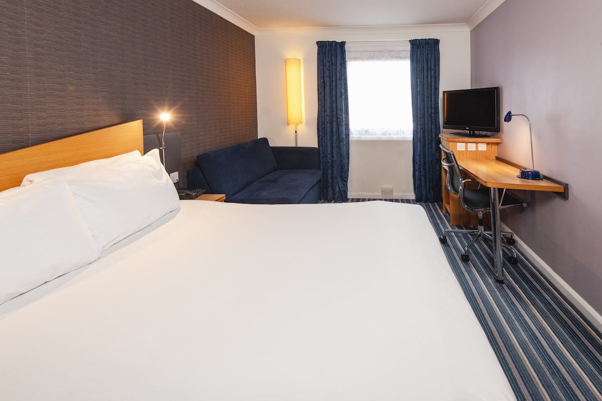 A hotel room with a large bed, a desk with a computer and TV, a lamp, and a window with curtains.