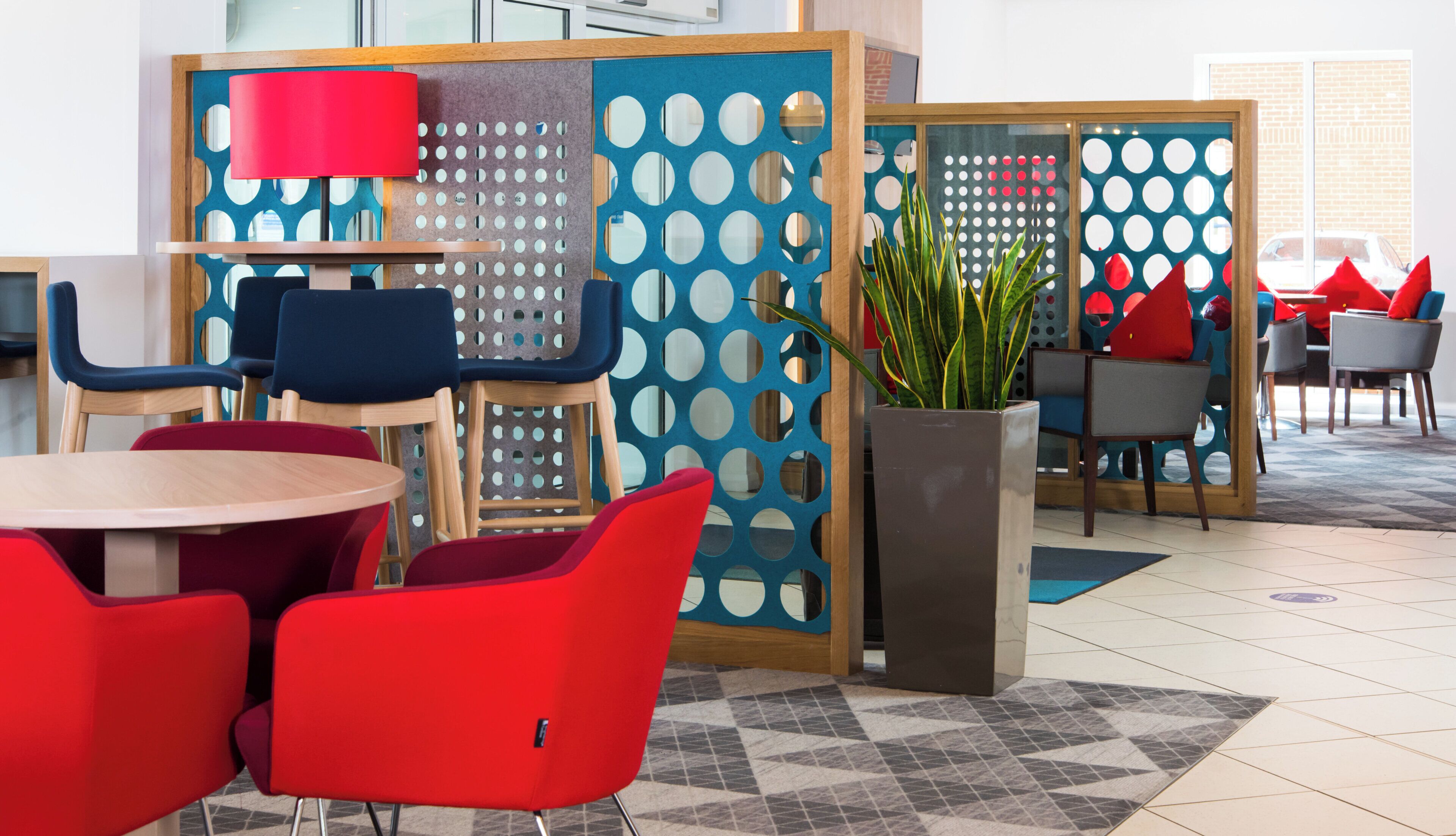 Een modern zithoek met ronde tafels en rode stoelen, een blauwe scheidingswand met ronde uitsparingen, en een potplant.