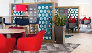 A modern indoor seating area with round tables and red chairs, a blue partition with circular cutouts, and a potted plant. - Holiday Inn Express Southampton - West by IHG (Southampton)