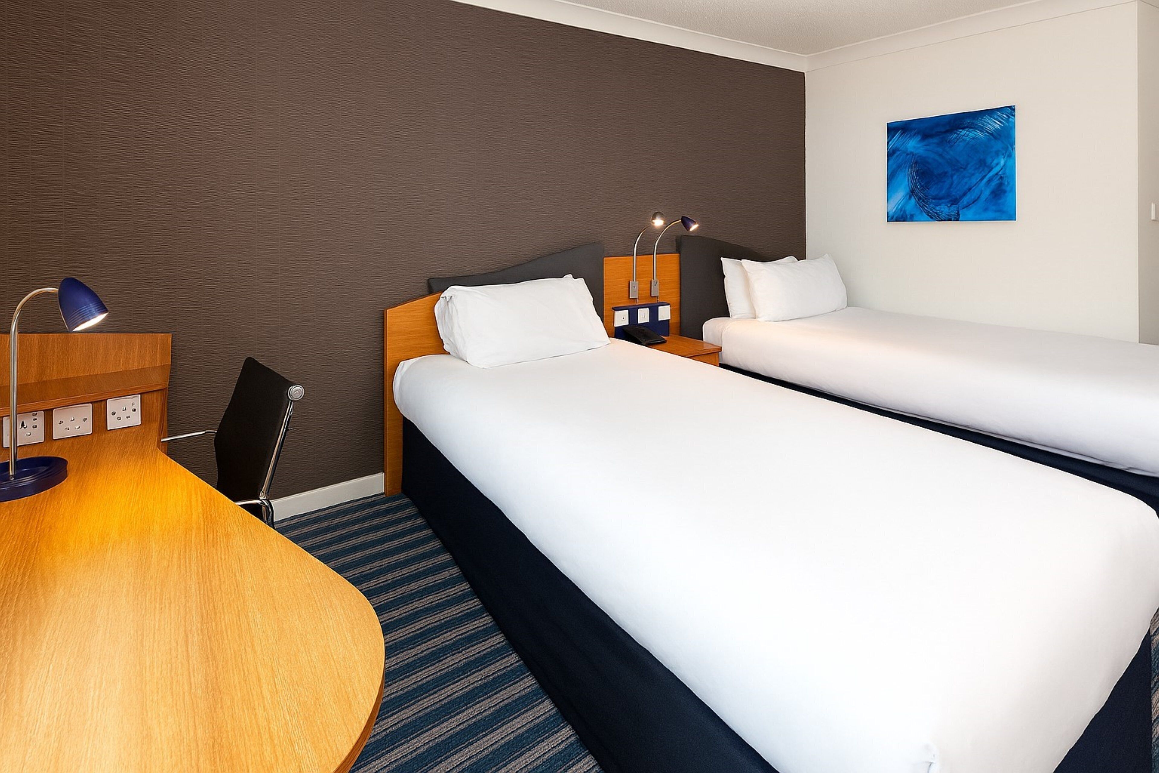 Een hotelkamer met twee bedden, een bureau, een lamp en een blauw abstract schilderij aan de muur.