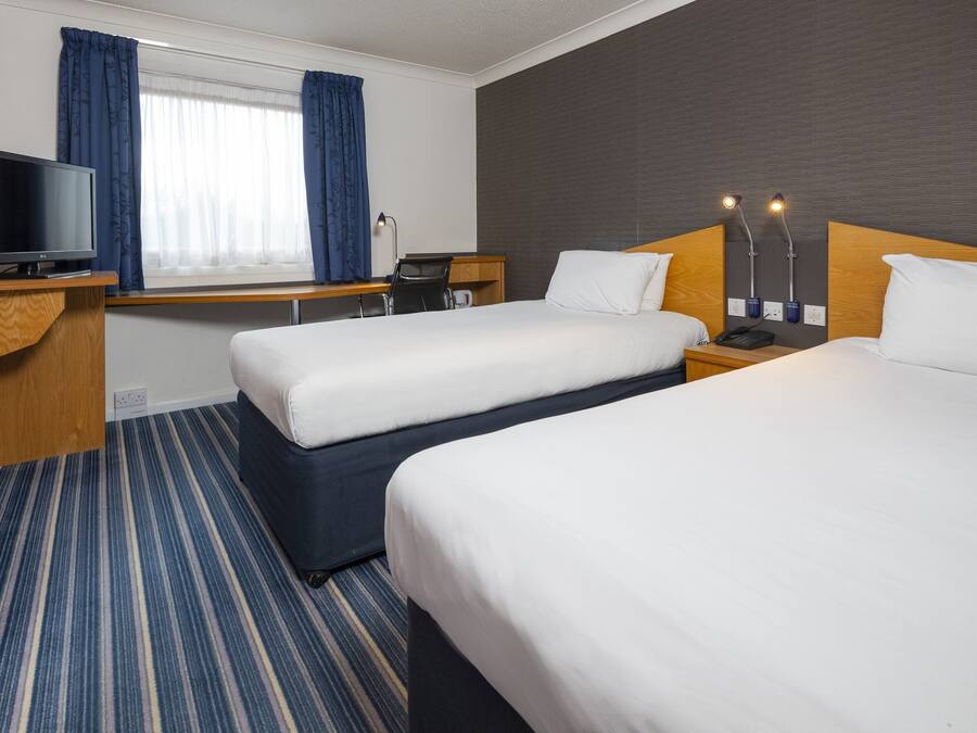 A hotel room with two beds, a desk with a TV, a chair, a lamp, and a window with curtains.