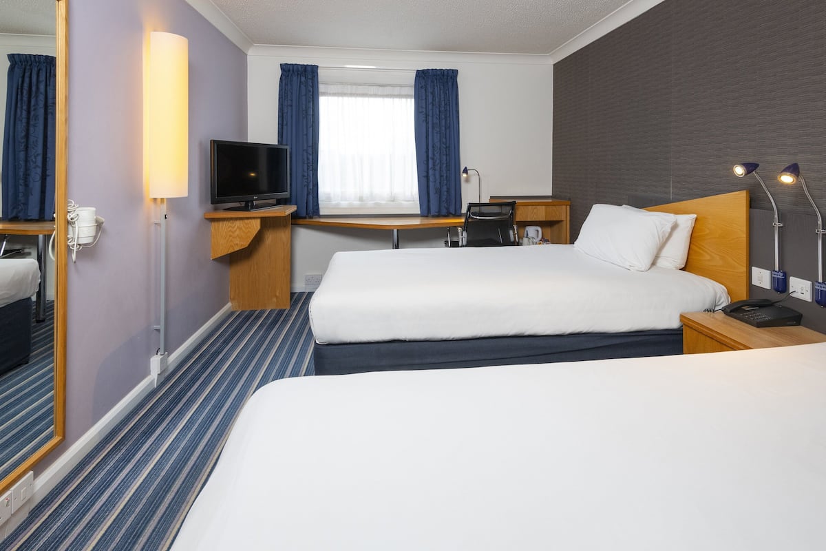 A hotel room with two beds, a desk, a television, and a window with curtains.
