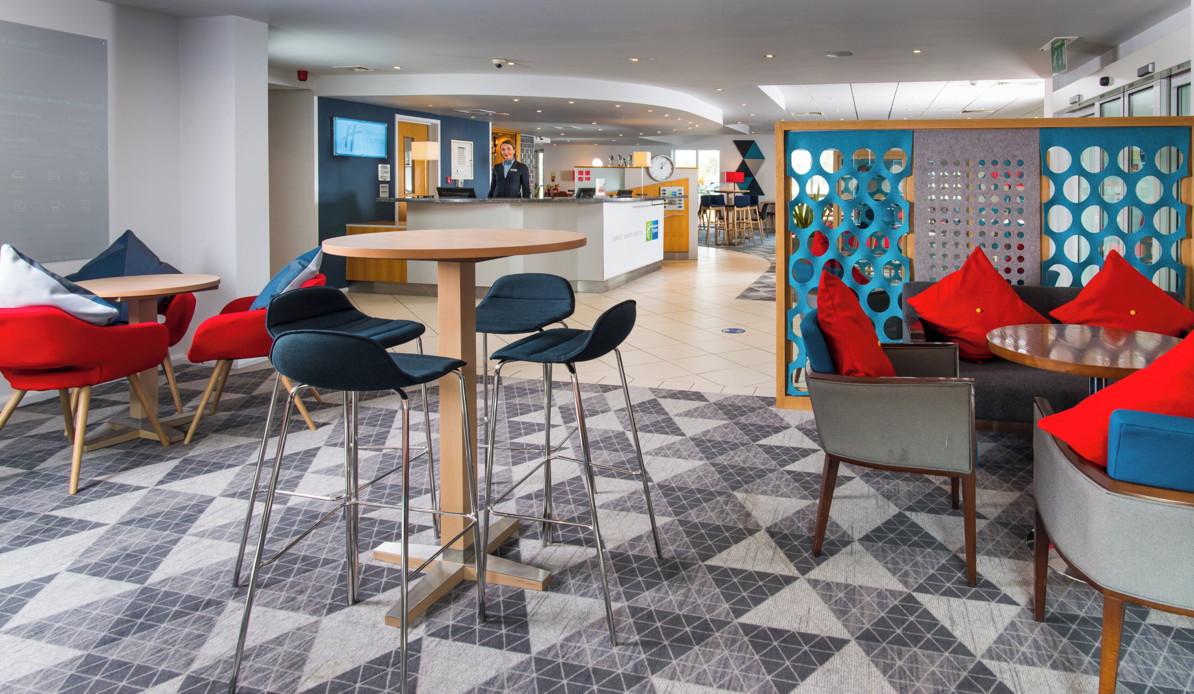 Un hall d’hôtel moderne comprenant un comptoir d’accueil, un espace salon avec des tabourets et des chaises de bar, et un sol à motifs.