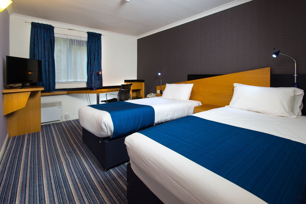 Ett hotellrum med två sängar, ett skrivbord, en stol, en tv och ett fönster med gardiner.