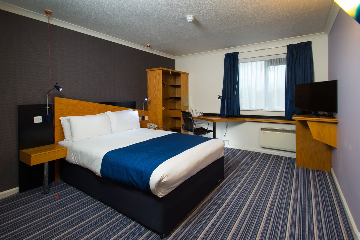 Ett hotellrum med en stor säng, ett skrivbord med en dator, en tv, en garderob och ett fönster med gardiner.