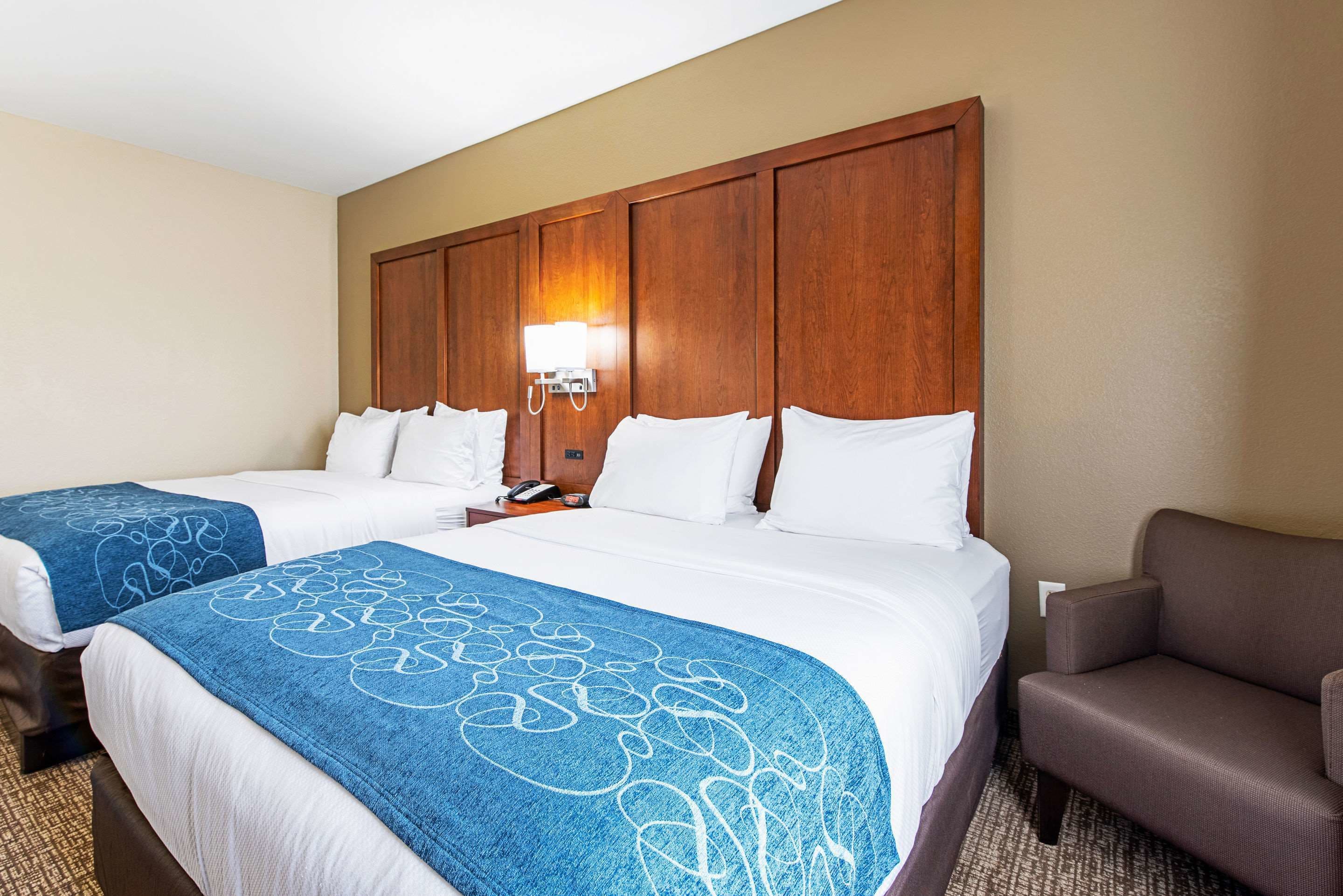A hotel room with two beds, a wooden headboard, a nightstand with a lamp, and a brown armchair.