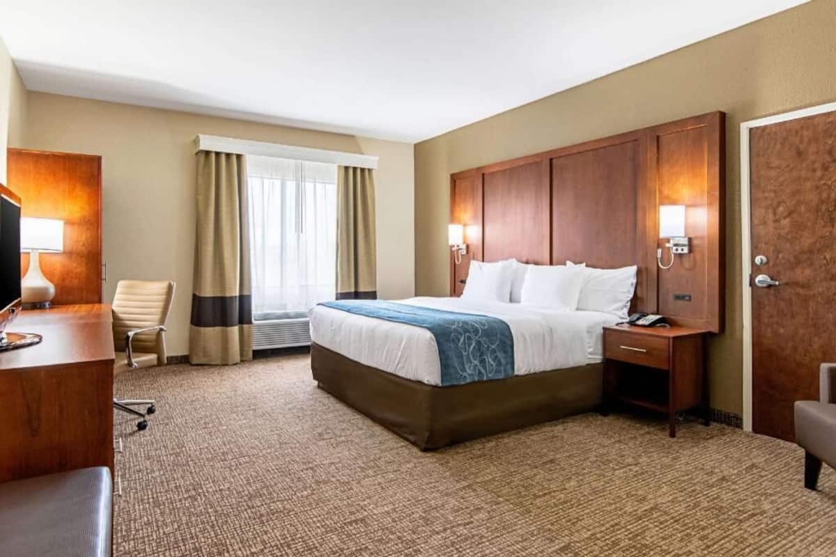 Ein Hotelzimmer mit einem großen Bett, einem Schreibtisch mit Lampe, einem Sessel, einem Fenster mit Vorhängen und einer Tür.