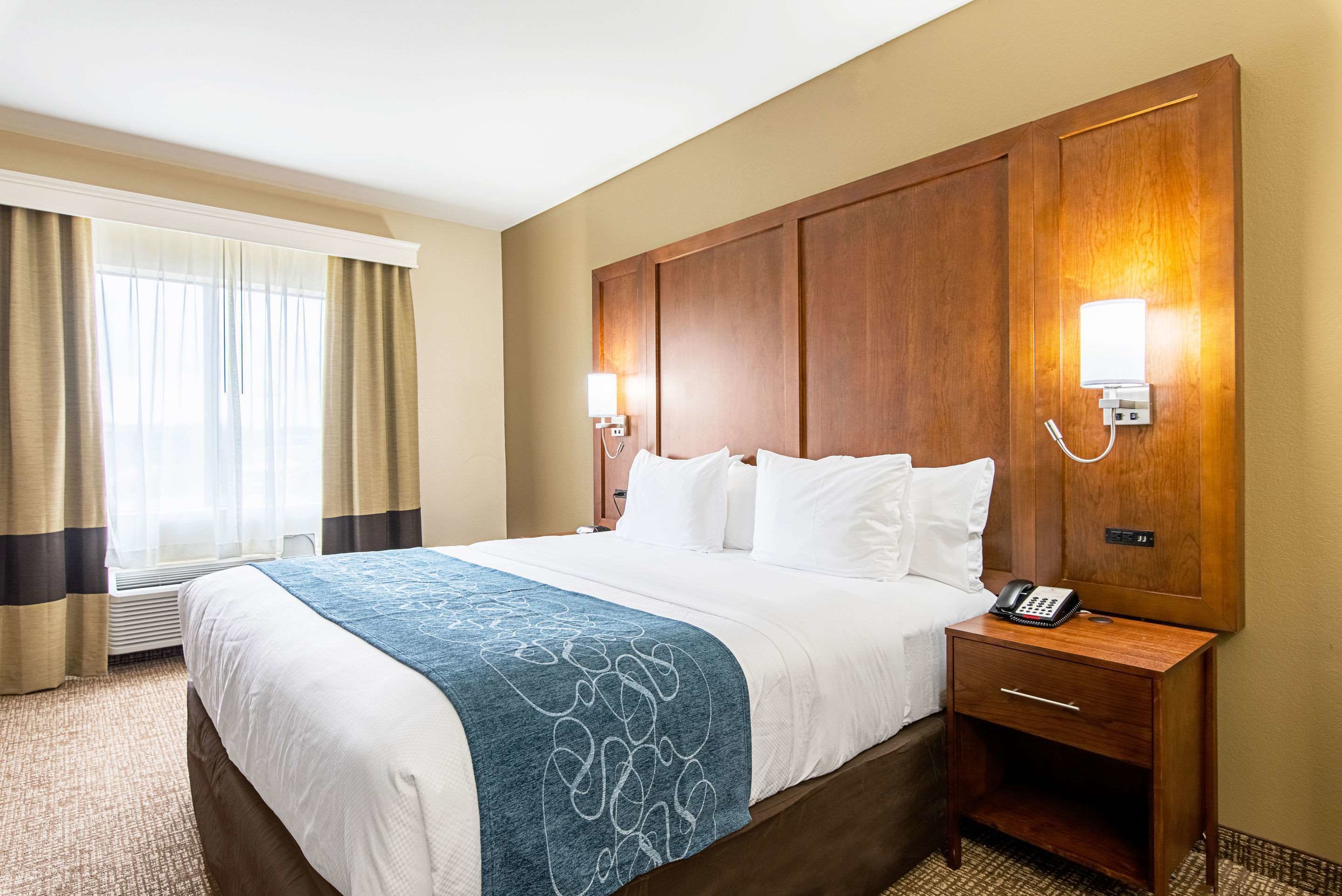 A hotel room with a large bed, a wooden headboard, two bedside lamps, a telephone, and a window with curtains.