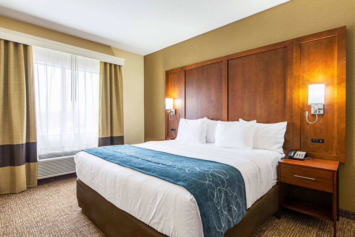 Ein Hotelzimmer mit einem großen Bett, einem Holzkopfteil, zwei Nachttischlampen, einem Nachttisch und einem Fenster mit Vorhängen.