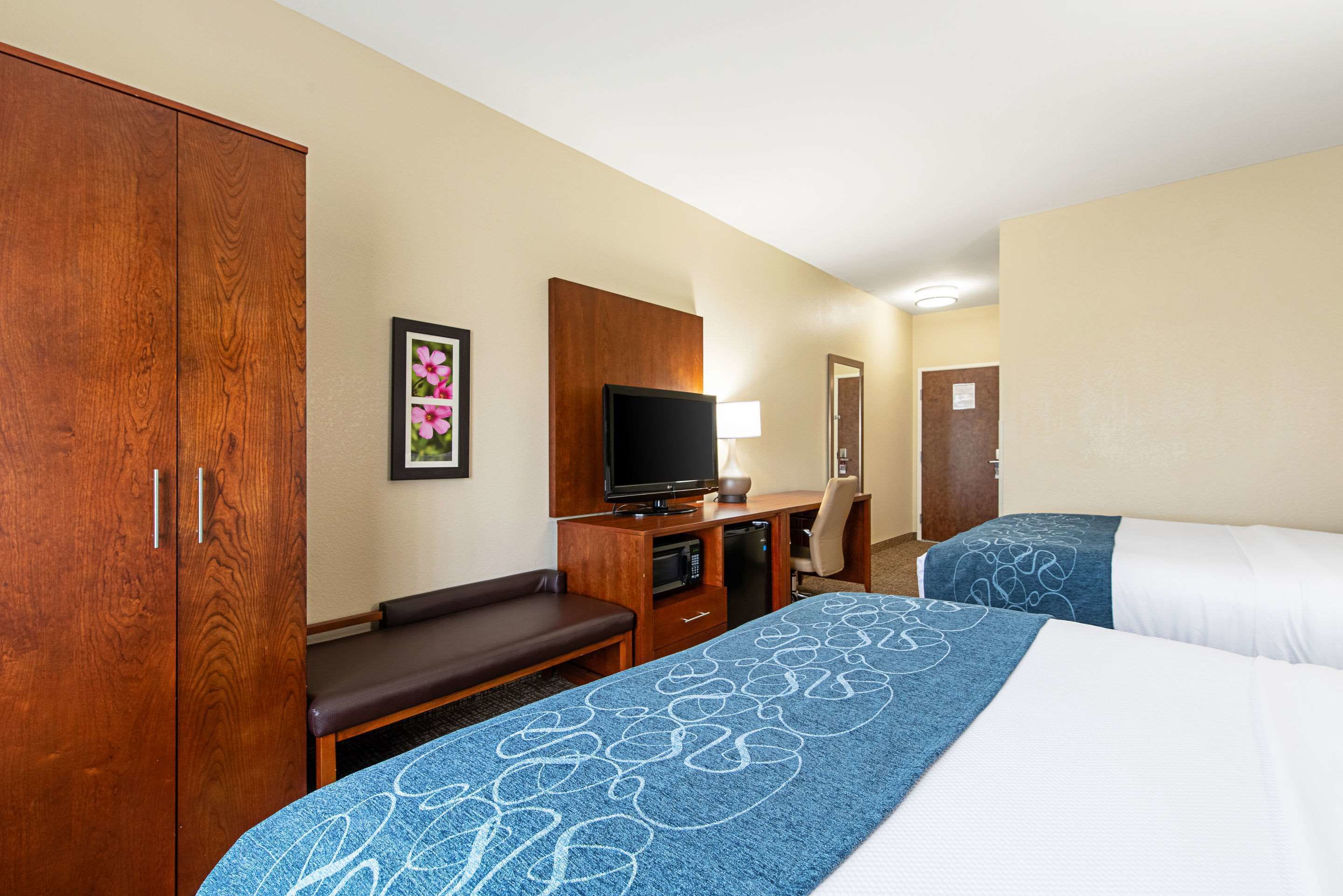 Habitación de hotel con un armario grande, un televisor, un escritorio con lámpara y dos camas con ropa de cama azul y blanca.