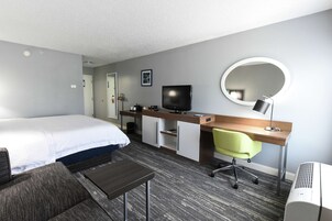 A hotel room with a bed, desk, chair, TV, and a round mirror.