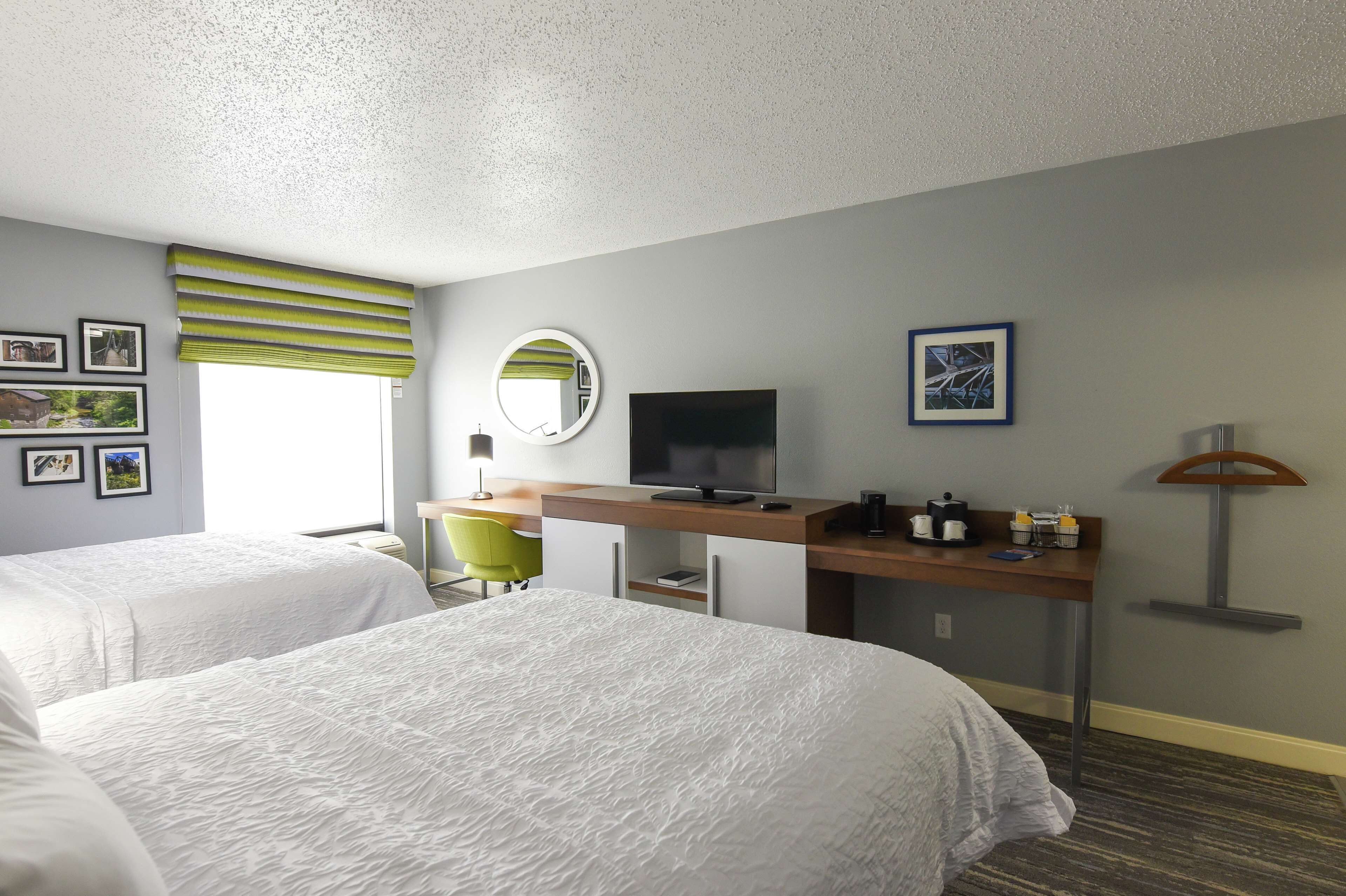 A hotel room with two beds, a desk with a TV, a mirror, and a window with blinds.