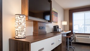 Habitación de hotel con televisión de pantalla plana sobre un escritorio de madera, cafetera, una silla y ventana con unidad de aire acondicionado.