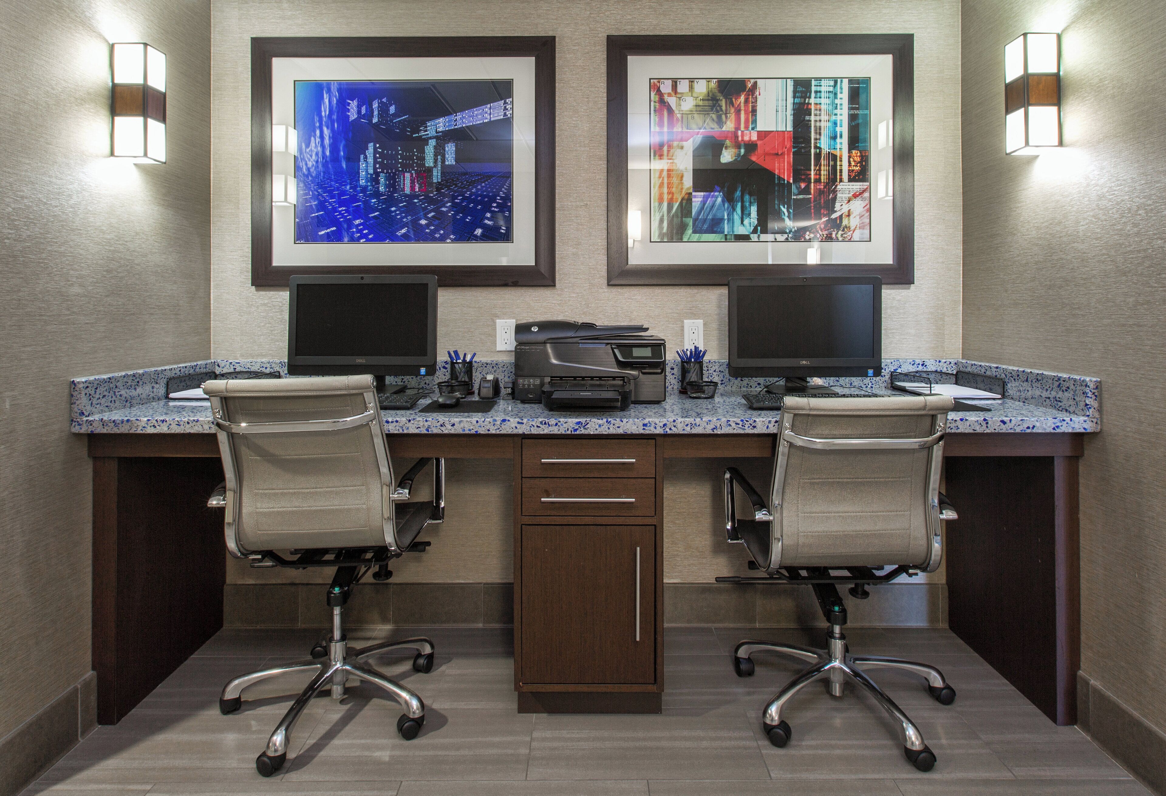 A hotel office with two desks, two chairs, two monitors, and framed artwork on the wall.