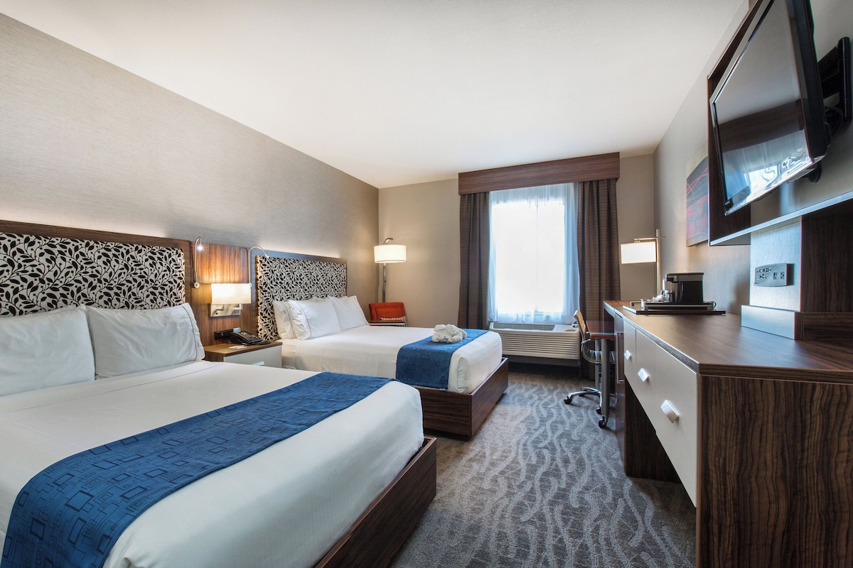 Quarto de hotel com duas camas, uma escrivaninha, uma televisão e uma janela com cortinas.