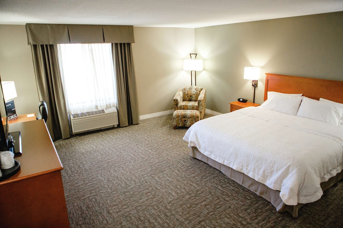 A hotel room with a large bed, a chair, a desk with a lamp, and a window with curtains.