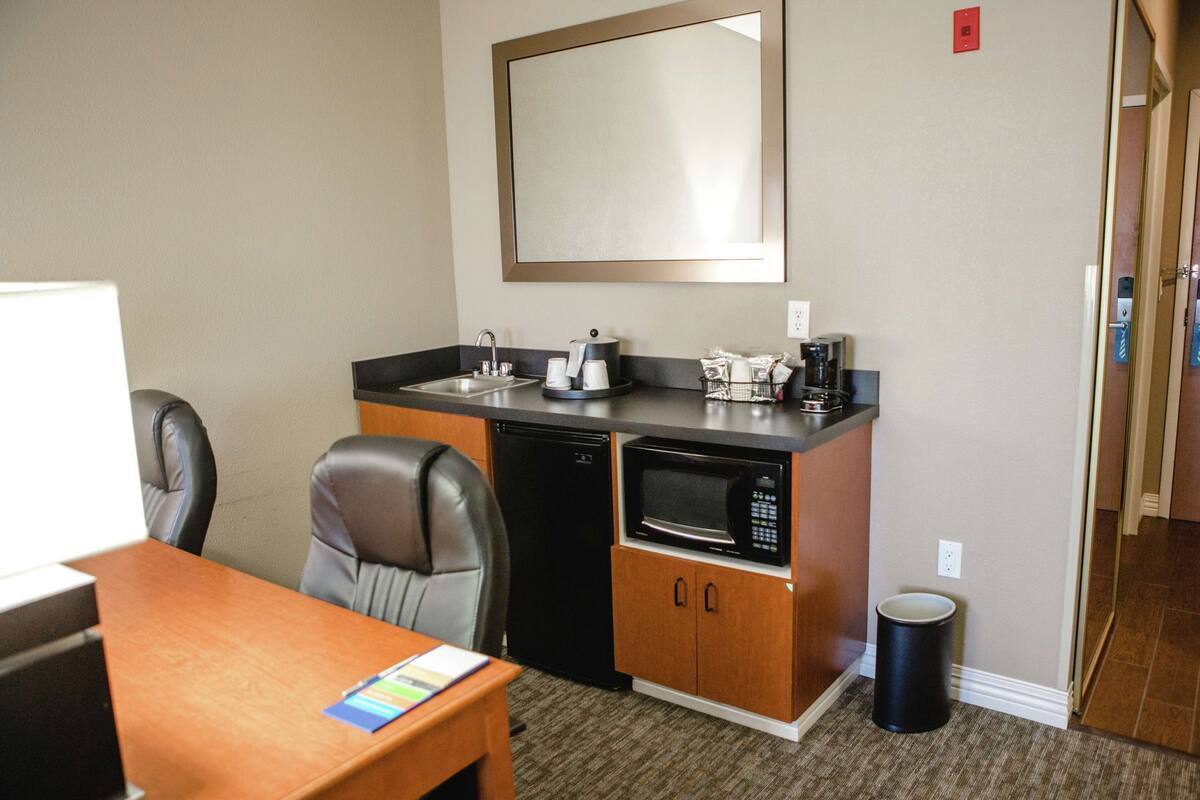 A compact kitchenette with a microwave, coffee maker, and sink. There is a trash can and a chair.