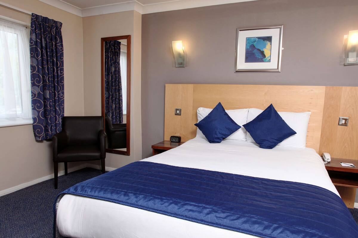 Une chambre d’hôtel avec un grand lit, deux oreillers, une chaise, une table de chevet, une horloge murale et un tableau encadré accroché au mur.