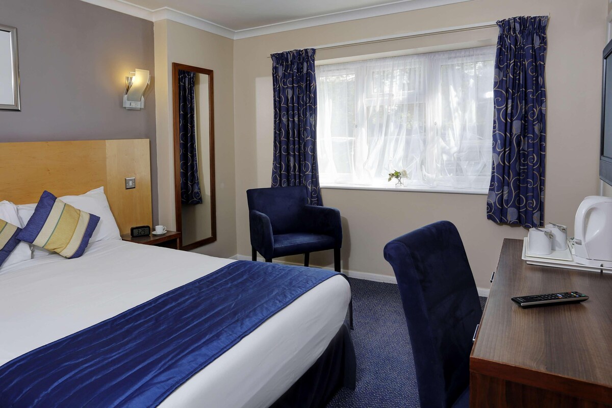 Een hotelkamer met een groot bed, een bureau met stoel, een klein tafeltje met waterkoker en gordijnen.
