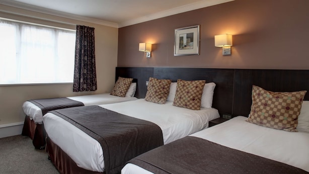 Une chambre d’hôtel avec trois lits, chacun recouvert d’une couverture marron et doté de draps blancs. Deux lampes murales et un tableau encadré sont accrochés au mur, au-dessus des lits.