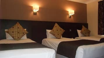 A hotel room with three beds, each with a dark headboard and white bedding. There are two wall-mounted light fixtures on the ceiling.