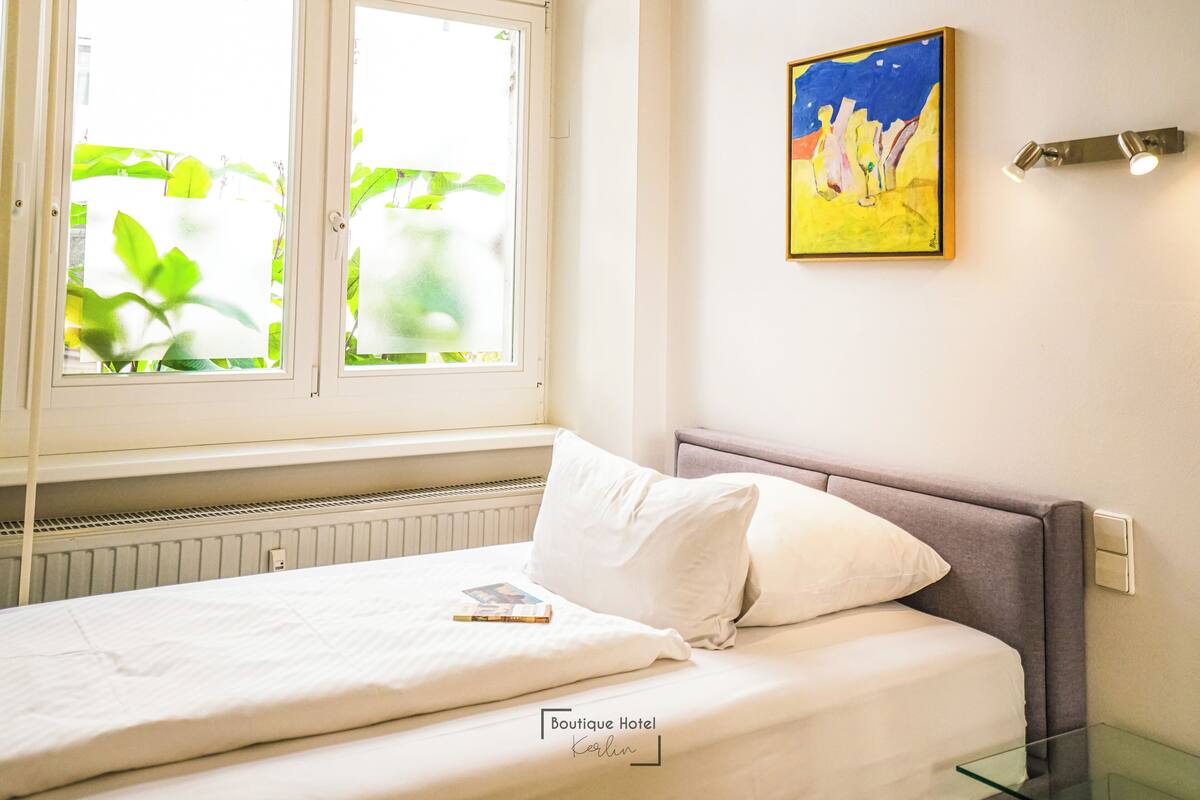 Ein Hotelzimmer mit Bett, Fenster mit Jalousien, einem Bild an der Wand und einem Nachttisch.