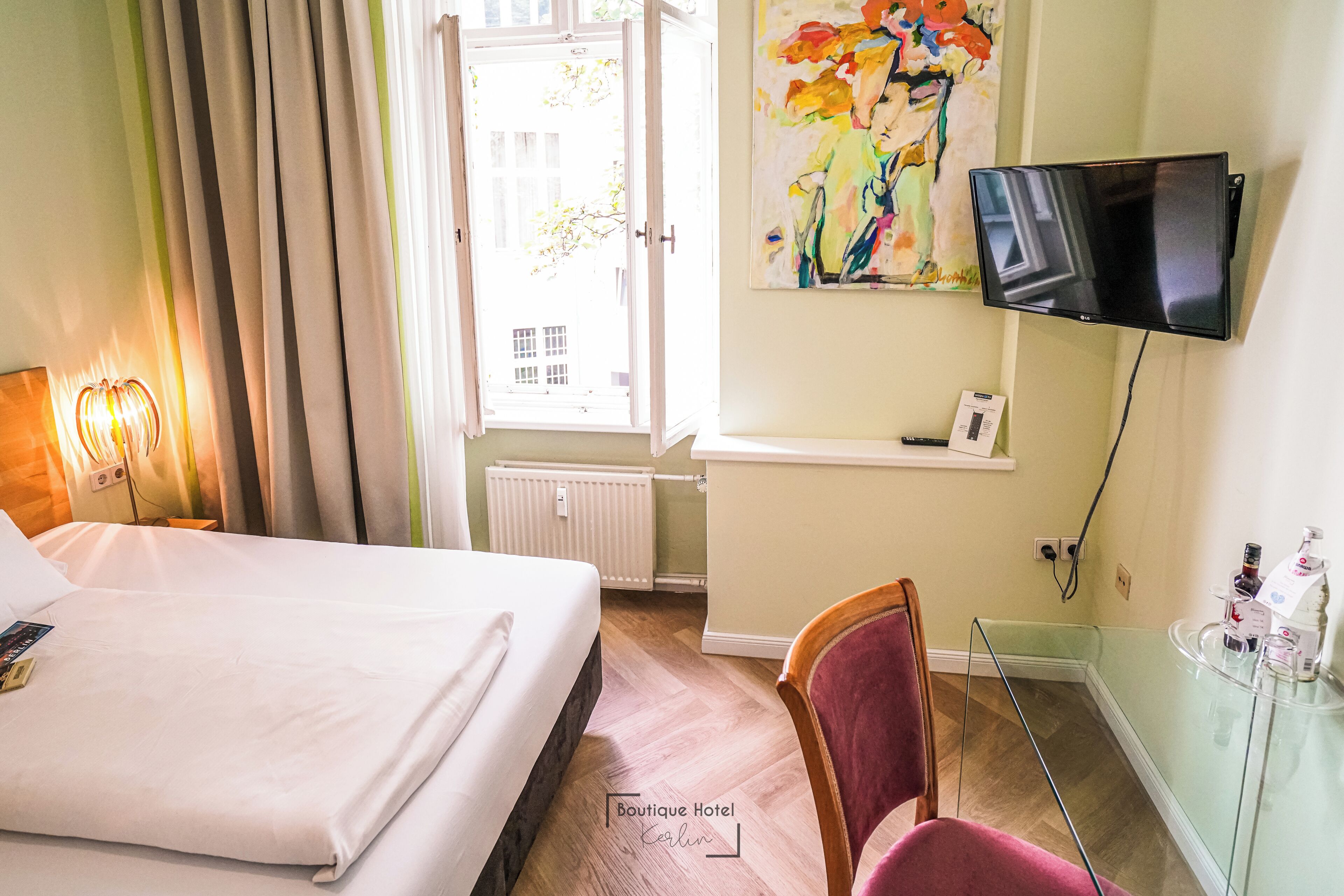 A hotel room with a bed, a chair, a TV, a window with curtains, and a painting on the wall.