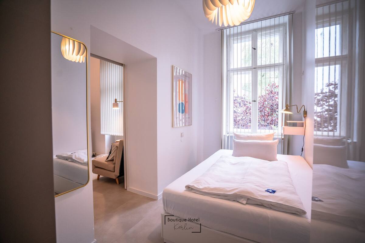 Ein Hotelzimmer mit einem großen Bett, einem Sessel, einem Schreibtisch und einem Fenster mit Jalousien.