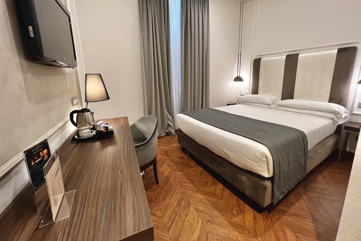 A hotel room with a bed, a desk with a lamp, a chair, and a TV mounted on the wall.