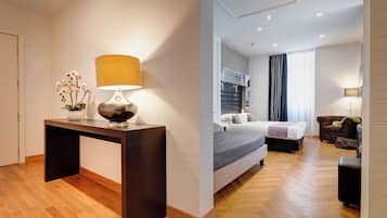 A modern hotel room with a wooden floor, a bed with a grey headboard, a bedside table with a lamp, and a small seating area with a sofa and armchair.